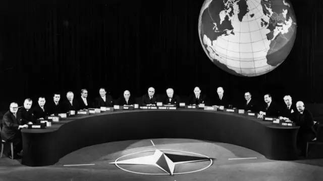 Delegates sit at U-shaped table at the Nato conference at Paris in a black and white photo, with the symbol of a globe behind them. From left, Van Acker (Belgium), Dieffenbaker (Canada), Gaillard (France), Adenauer (Germany), Hansen (Denmark), Karamanlis (Greece), Jonasson (Iceland), Zoli (Italy), Spaak (NATO Secretary-General), Bech (Honorary Chairman), Hommel (Luxembourg), Luns (Netherlands), Gerhardsen (Norway), Cunha (Portugal), Menderes (Turkey), Harold MacMillan (Britain) and Dwight D Eisenhower (USA)