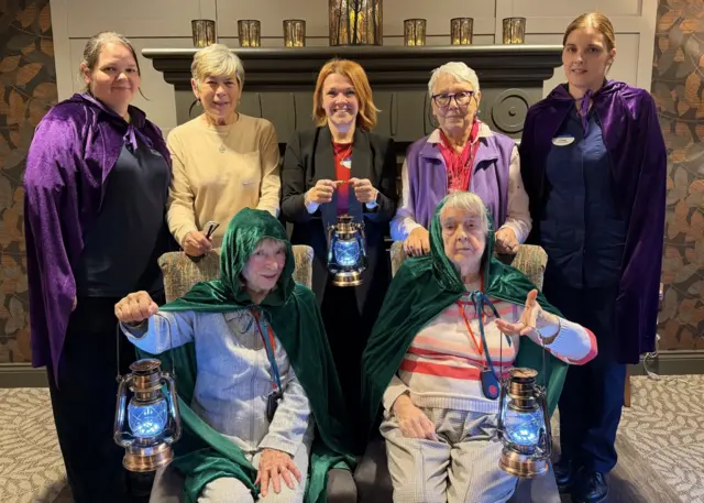 Se puede ver a siete miembros del personal y residentes de la residencia de ancianos Parkfield Grange con capas azules y verdes. Tres de ellos sostienen lámparas al estilo del programa de televisión Traidores.