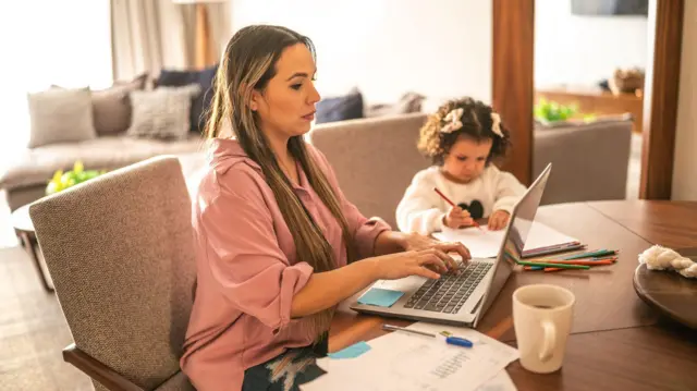 Uma mãe trabalha no computador enquanto a filha pinta