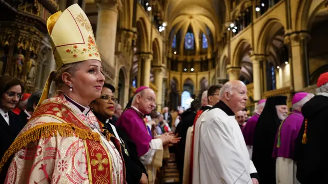 Members of di clergy stand and wait inside di cathedral