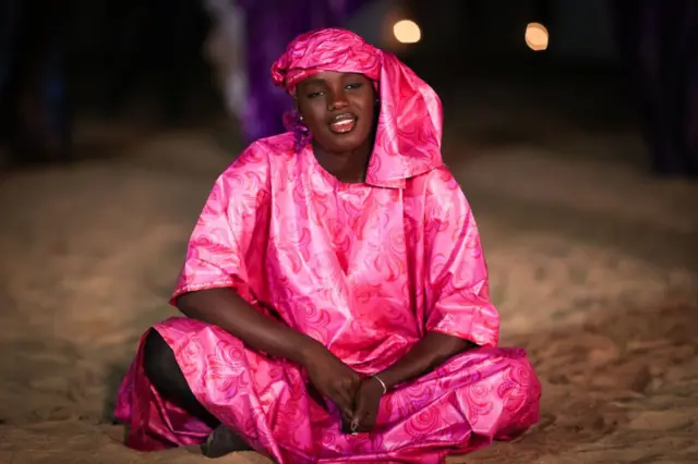 A woman sits cross-legged on the ground and sings.