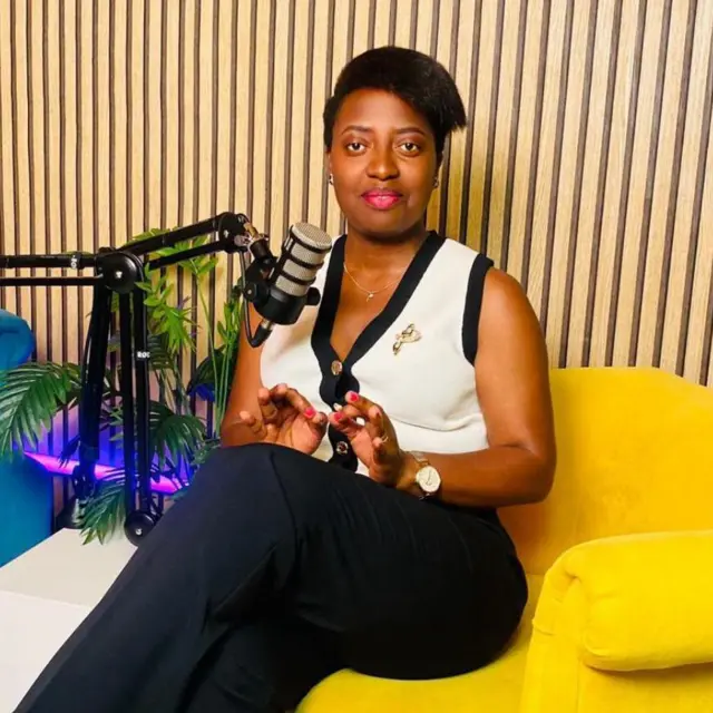 Marlène aba Hambourg mu Budagi yambaye ishati y'umweru n'ipantaro y'umukara, yicaye ku ntebe y'umuhondo kandi imbere ye hari microphone