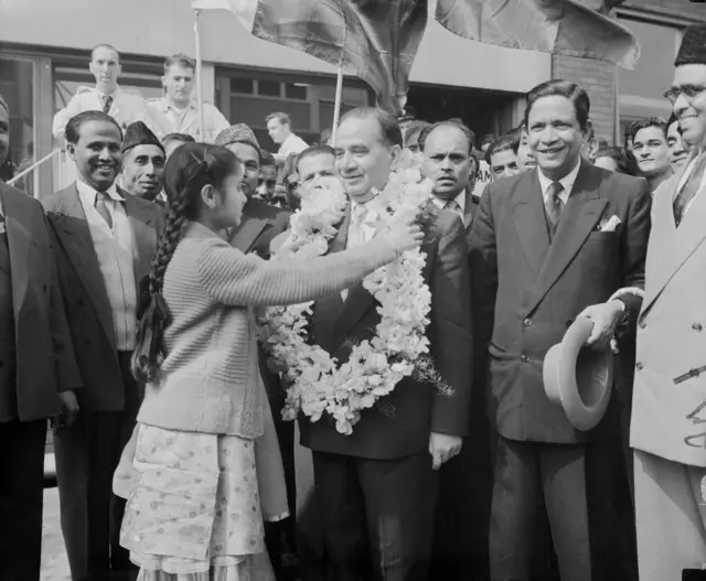 সাদাকালো ছবিতে হোসেন শহীদ সোহরাওয়ার্দীর গলায় মালা পরাচ্ছে একটি কিশোরী। পাশে কোট-প্যান্ট পরা কয়েকজন হাঁসিমুখে দাঁড়িয়ে আছেন