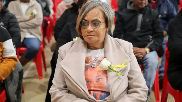 Amanda Smith-Daniels, grandmother of Joshlin Smith, wear one beige jacket on top T-shirt wey dem print wit Joshlin foto. One white rose dey pinned to her lapel. She siddon on top one red plastic chair wit odas wey dey wait for di sentencing hearing to start for di Western Cape High Court in Saldanha Bay - 29 May 2025.