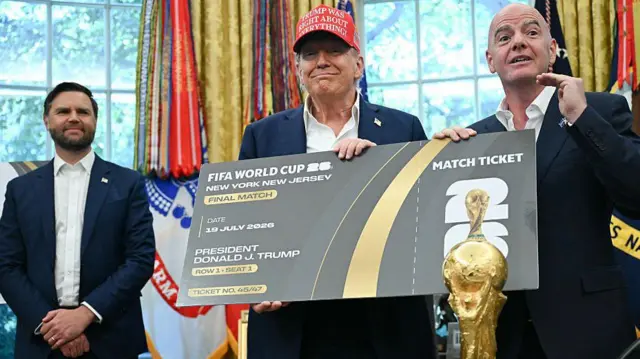Zdjęcie zrobione w Gabinecie Owalnym w Waszyngtonie, USA. Prezydent USA Donald Trump i prezydent FIFA, Gianni Infantino, trzymają bilet na finał Mistrzostw Świata w 2026 r. Trump ma na sobie czerwoną czapkę z wyszytym z przodu napisem ,,Trump was right about everything" (,,Trump miał rację co do wszystkiego.") W tle wiceprezydent USA, JD Vance. Na pierwszym planie widoczne biurko, na którym stoi Puchar Świata