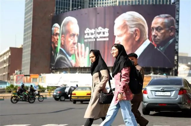 A billboard at a traffic intersection in Tehran shows the Iranian president and a Revolutionary Guards leader opposite Biden and Netanyahu. Three people walk in front of the billboard