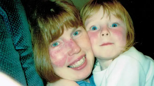 Жінка з блакитними очима та рудим волоссям тримає на руках дівчинку з блакитними очима та рудим волоссям. Вони сидять на зеленому дивані. 