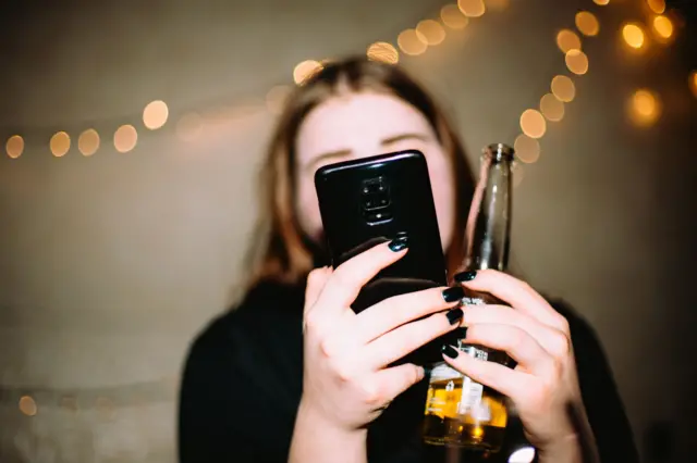 Молода жінка тримає в одній руці смартфон, а в іншій відкриту пляшку з пивом. На задньому плані видно розмиті теплі вогники гірлянди.