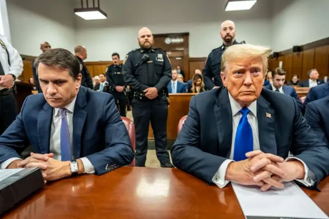 Тодд Бланш и Дональд Трамп, оба в костюмах с синими галстуками, опираются руками на стол в зале суда в Нью-Йорке.