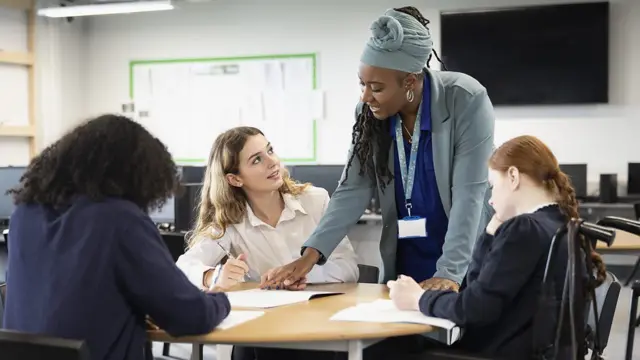 Um grupo de mulheres de diferentes etnias senta em uma mesa, enquanto uma mulher negra, de pé, aponta algo em um caderno; ela parece ser a professora, pois o grupo está em sala de aula