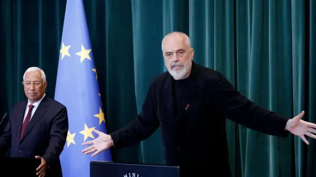 edi rama u crnoj majici i pantalonama, iza njega sa leve strane sedi muškarac u odelu i kravati