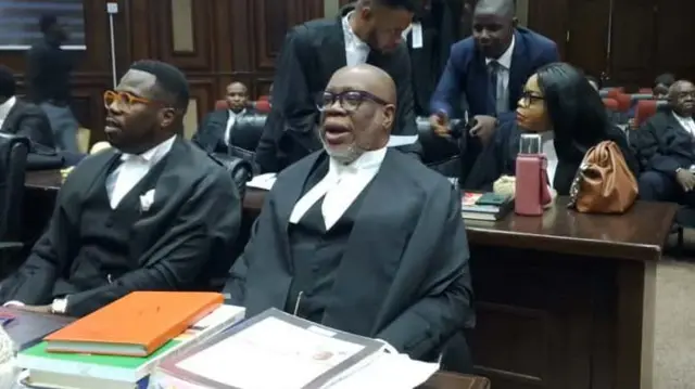 Okaịwụ mmadụ abụọ nọ na-ele anya dịka ndị okaịwu ndị ọzọ nọ ha n'azụ. Akwụkwọ dị iche iche nọ ha n'ihu n'elu tebụlụ.