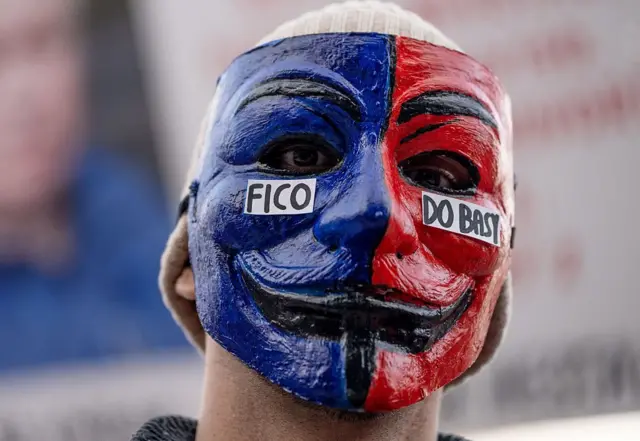 Синьо-червона маска Гая Фокса на обличчі протестувальника, через яку видно лише очі, що дивляться у камеру.