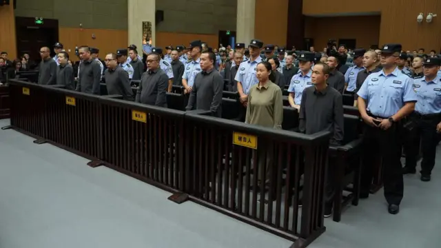 Seven men and a woman, dressed in grey and beige shirts stand in a row in a court, surrounded by security officers in blue shirts with black caps