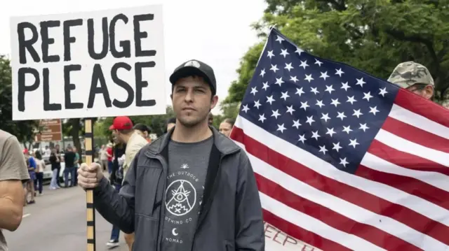 One White South African carry placard wey dem write "Refuge Please" ontop as e stand behine US flag.