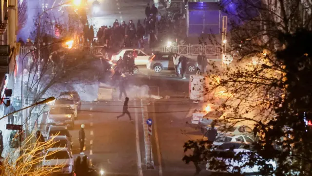 Nocna scena uliczna przedstawiająca protestujących gromadzących się wokół zaparkowanych samochodów. Widoczny jest dym, a w pobliżu tłumu płoną ogniska.
