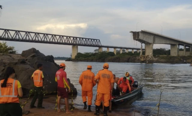 Bombeiros fazem buscas por desaparecidos com barco. No fundo está a ponte que desabou.