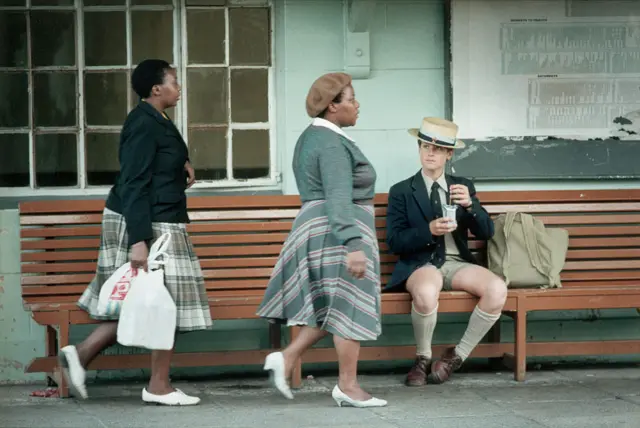 African domestic workers catching a train to Crossroads, a camp on the outskirts of Cape Town, pass by a white student from Bishops, an exclusive Cape Town prep school, who is also waiting for a train.
