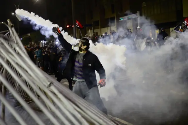 Протестующий в противогазе и перчатках поднимает правую руку, стоя среди слезоточивого газа рядом за частично разрушенным металлическим ограждением. За ним собралась группа людей.