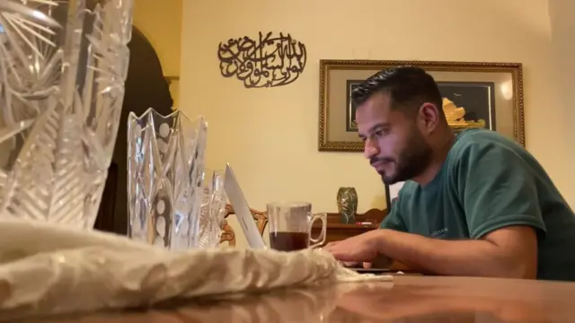 Amir Azaizeh tape sur son ordinateur portable dans une pièce. Il est penché en avant. Une tasse en verre contenant une boisson foncée est à portée de main. Des ornements en verre et un tissu couleur crème sont au premier plan.