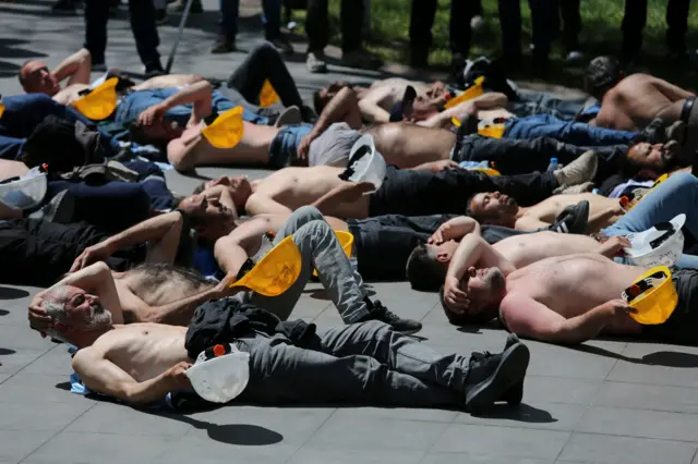 Kurtuluş Parkı'nda açlık grevi yapan madenciler üstleri çıplak bir şekilde yere uzanmış, sağ ellerinde sarı baretlerini tutuyor. 