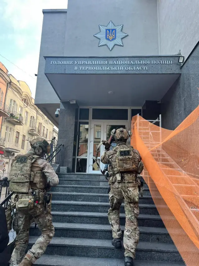 Біля входу до будівлі з написом «Головне управління Національної поліції в Тернопільській області» двоє озброєних силовиків у формі піднімаються сходами. На їхніх спинах видно нашивки «СБУ», вони тримають зброю та рухаються до дверей.