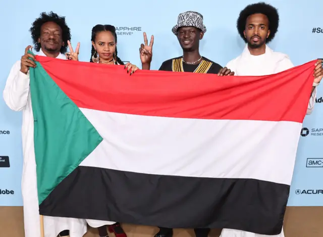 Four people stand in a row at a photocall, holding up the Sudanese flag.