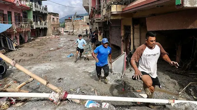 Nepal'de ani selin ardından hasar gören binalar arasında çamur ve molozla kaplı bir sokakta üç adam yürüyor.