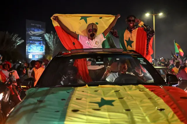 Les supporters sénégalais célèbrent leur victoire sur la Place du Souvenir Africain à Dakar. Un fan portant un masque de lutteur mexicain brandit un drapeau sénégalais et se tient debout sur le toit d'une voiture, dont le capot est également orné du drapeau.