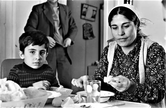 Bir anne ve oğlu masada yemek yiyor. Arkada babaları olduğunu düşündüğümüz bir adam ayakta duruyor. 