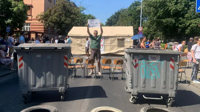 Muškarac stoji sa transparentom na sred ulice okružen kontejnerima tokom blokade