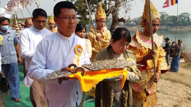 ဘုရားကြီးရွာမှာ ၂၀၂၃ ခုနှစ်၊ မတ်လတုန်းက ပြုလုပ်ခဲ့တဲ့ ထီးတင်ပွဲမှာမြင်ရတဲ့ စစ်ကောင်စီ ဧရာဝတီတိုင်း ဝန်ကြီးချုပ် ဒုတိယဗိုလ်ချုပ်ကြီးဟောင်း ဦးတင်မောင်ဝင်း၊ ဓာတ်ပုံ ညာဘက် မျက်မှန်တပ် တိုက်ပုံဝတ်အမျိုးသားတစ်ဦးနဲ့ ပဝါခြုံထားတဲ့အမျိုးသမီးတစ်ဦး အဝါရောင်ပိတ်စ ခင်းထားတဲ့ ဗန်းနဲ့ ငှက်မြတ်နားတော်ကို သယ်လာတာတွေေ့ရ၊ ဒေဝါဝင်း သရုပ်ဖော် အဝါရောင် နတ်ဝတ်စုံ ၀တ်ထားသူ အမျိုးသားအချို့လည်း မြင်ရ၊ ဓာတ်ပုံ အနောက်ဘက်မှာ မြစ်ပြင်ကျယ်နဲ့ သာသနာ့အလံတိုင်ကို မြင်ရ 
