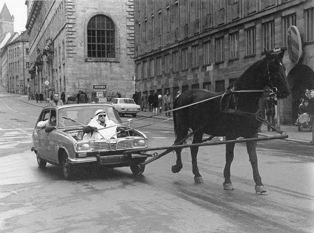 عکس سیاه و سفید، مردی در لباس عربی به جای موتور خودرویی نشسته و افسار اسبی که در حال کشیدن اوست، در دست دارد