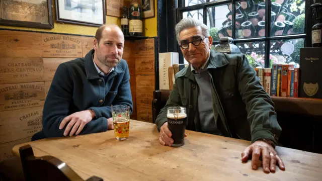 Принц Уильям и Юджин Леви сидят за деревянным столом в пабе. Перед Уильямом стакан с сидром, у Леви в руке пинта темного пива.