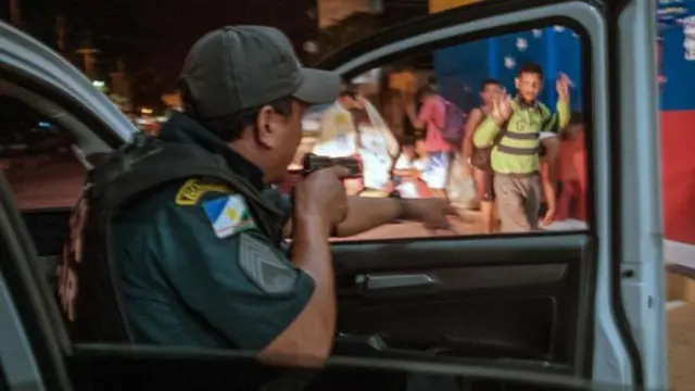 Policial abrindo porta de viatura com a câmera apontada, enquanto homem ao lado ergue as mãos para demonstrar que está desarmado