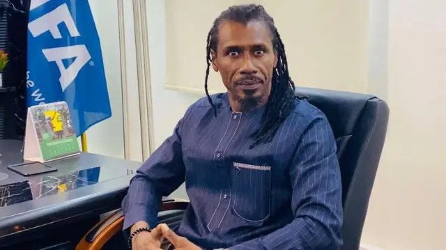 Aliou Cissé porte une chemise à rayures bleu foncé alors qu'il est assis sur une chaise de bureau avec des murs blancs derrière lui et un bureau et un drapeau bleu de la FIFA visibles à sa droite.