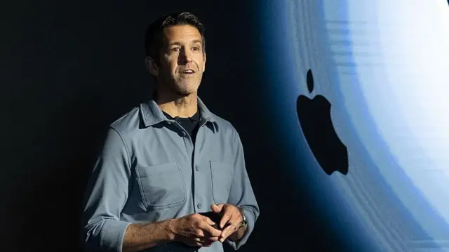 John Termus, con camisa gris, frente a una pantalla que proyecta el logotipo de Apple, durante la presentación de un producto.