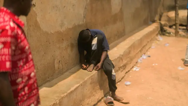 Un jeune homme est assis sur une corniche près d'un mur. Il est affaissé, de sorte qu'on ne voit pas son visage. 