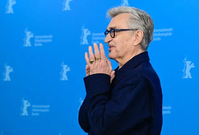 Wim Wenders arkada mavi Berlinale fonu önünde ellerini birbirine birleştirmiş şekilde görülüyor. 