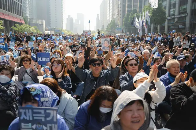 높고 현대적인 건물이 늘어선 시내 거리에 많은 이재명 지지자들이 모였고, 그중 일부는 파란색 플래카드를 들고 있다