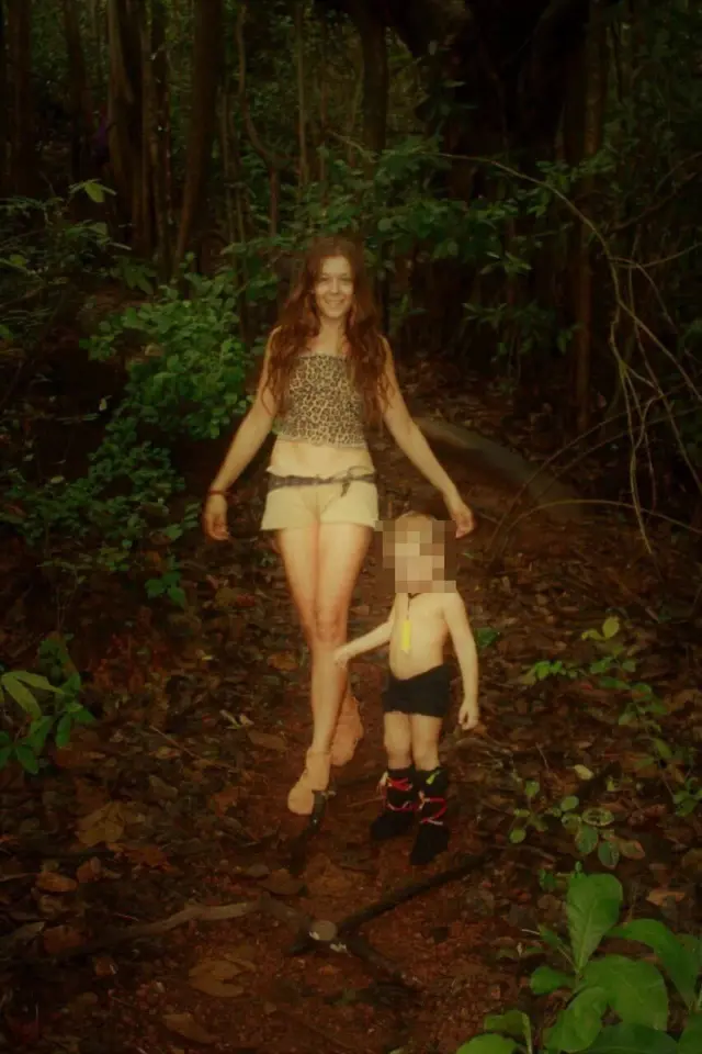 Улыбающаяся Нина Кутина с длинными темными волосами в шортах и майке леопардового цвета позирует на тропинке в джунглях с маленьким ребенком лет четырех