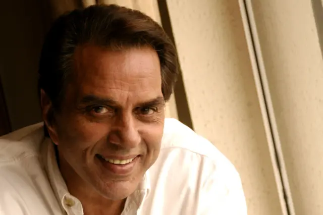Dharmendra wears a white button-up shirt, sitting indoors near a window.