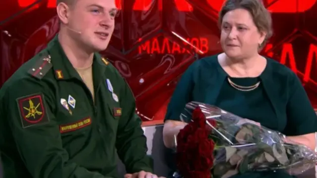 30 ёшлардаги ҳарбий кийимдаги эркак телевидение дастурида гапиряпти, стол устида ининг олдида бир даста қизил гуллар жойланган.