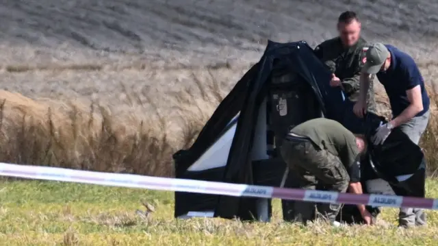 За огороджувальною стрічкою у полі двоє чоловіків у військовій формі та один у цивільному одязі огортають дрон чорним матеріалом, ймовірно, для транспортування