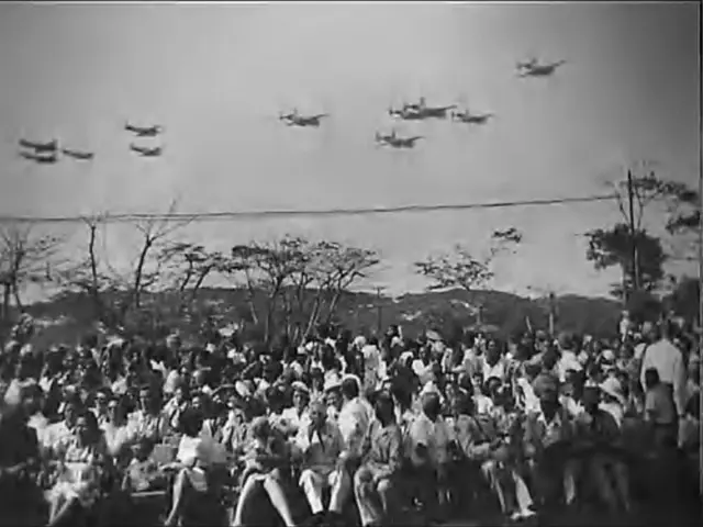 Parade militer di gelar di depan Istana Koningsplein, Batavia, untuk memperingati Hari Ratu. Acara itu dihadiri ribuan orang. Para pejabat dan orang Belanda berada di lingkaran utama parade, sedangkan warga lokal berada dari kejauhan.
