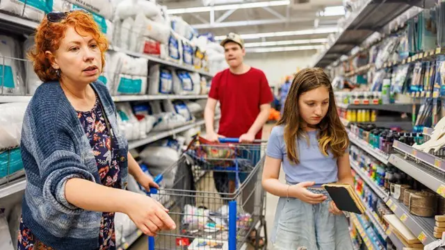 Casal com filha no supermercado. O pai empurra o carrinho, enquanto a filha escolhe um caderno e a mãe parece coordenar a situação