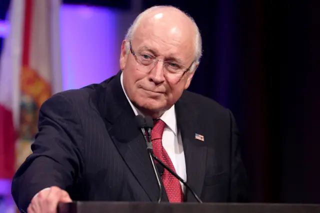 Dick Cheney is standing at a podium, wearing a dark pinstripe suit with a white dress shirt and a red patterned tie. A small American flag pin is visible on the left lapel. He is speaking into a microphone, and part of a flag with red and white stripes is visible in the background, along with purple stage lighting. The setting appears to be a formal event or conference.