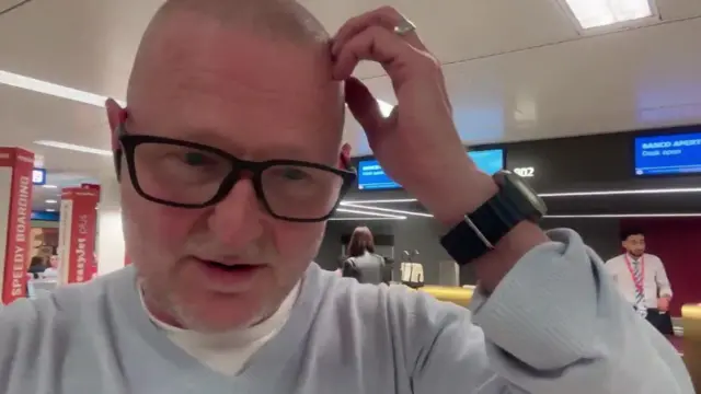 Mężczyzna stoi na lotnisku, trzymając dłoń na głowie. Ma na sobie luźny szary sweter i okulary w czarnych oprawkach