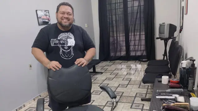 Lucas aparece em pé, vestindo uma camiseta preta, atrás de uma cadeira, com equipamentos de barbeiro ao lado