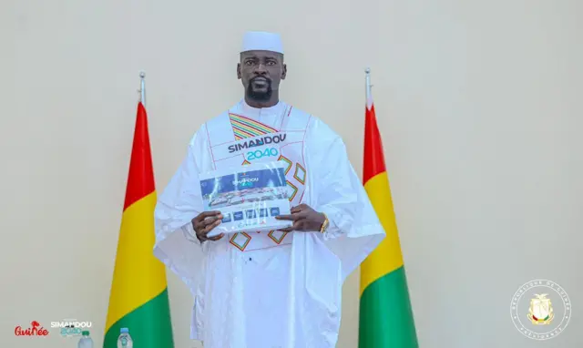 Le Général Mamadi DOUMBOUYA, ici en boubou blanc devant deux drapeaux guinéen, reçoit des documents du Programme de Développement Socio-Économique Durable et Responsable Simandou 2040.